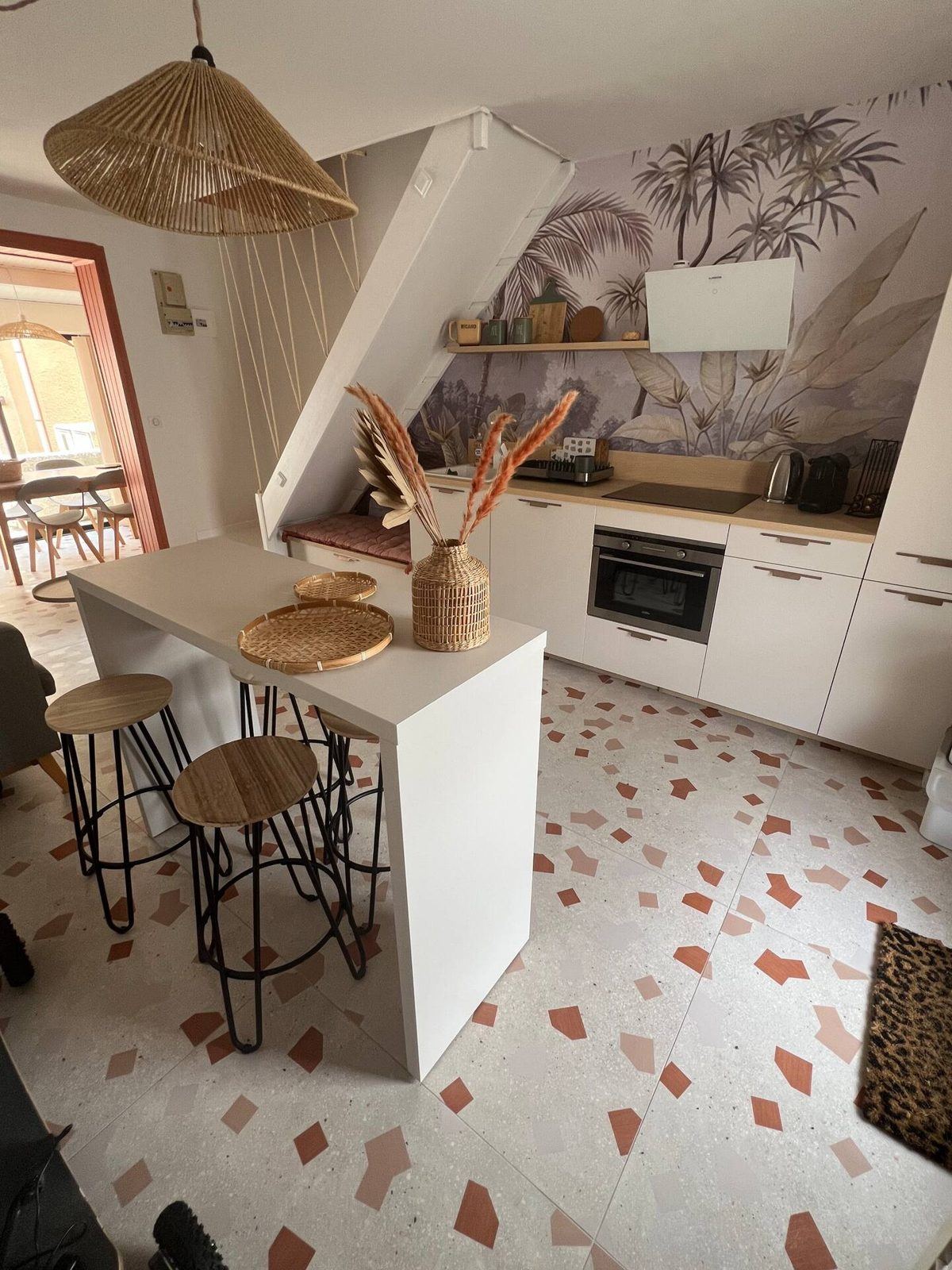 Pièce de vie de la location à Narbonne-Plage : îlot de cuisine, suspension en rotin, papier peint palmiers